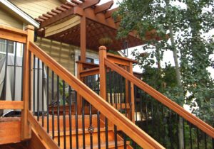 A beautiful redwood deck with Cedar pergola. The railing features metal balusters with a basket design