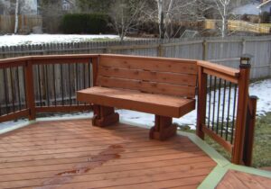 This redwood deck is laid out in a herringbone pattern with a single picture frame around it. The railing has wood components with black metal balusters and post cap lights at the stairs. We also custom-built a small bench to match the deck.