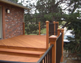 A redwood deck with the boards laid out at different angles on each level. The wood posts of the railing each have a post cap, and between the posts is a black, metal panel railing.