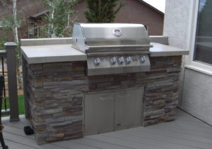 An outdoor cooking area built with a stone base, tiled countertop and backsplash, and a storage cabinet under the grill.